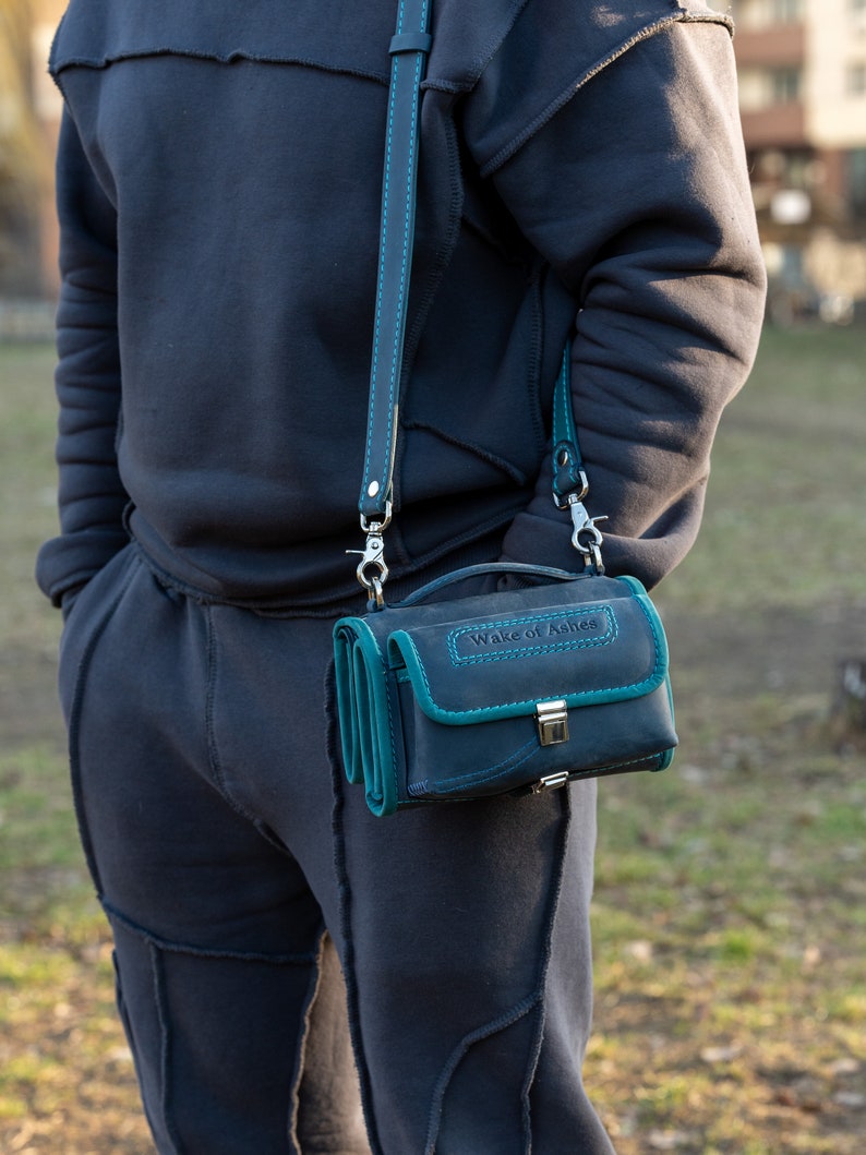 Puede incluir: Un bolso bandolera de cuero azul oscuro con ribetes y costuras turquesas. El bolso tiene un cierre de solapa y una correa desmontable. El texto "Wake of Ashes" est&aacute; grabado en la parte superior.