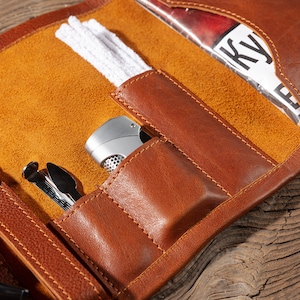May include: A brown leather tobacco pouch with multiple pockets and compartments. The pouch is open and shows a lighter, a pipe cleaner, and a pack of tobacco.