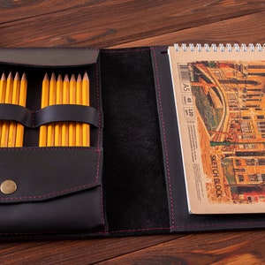May include: Black leather sketchbook case with a zipper closure and a built-in pencil holder. The case is open to reveal a sketchbook with a spiral binding and a drawing of a cityscape. The sketchbook is labeled "Sketch Block" and has the dimensions "A5 40 120".