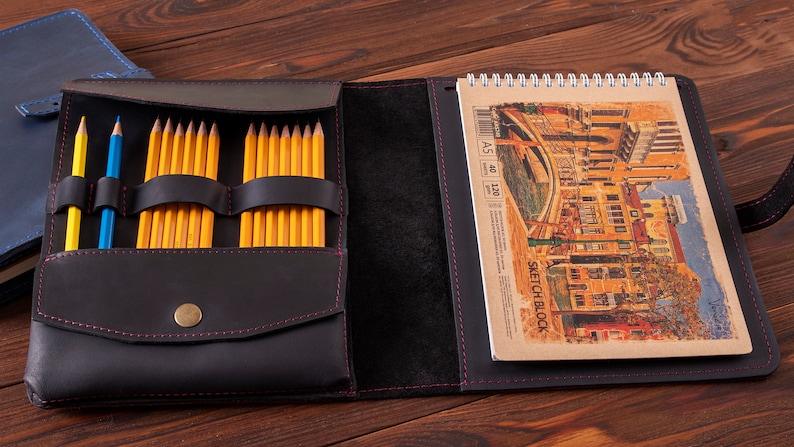May include: Black leather sketchbook case with a zipper closure and a built-in pencil holder. The case is open to reveal a sketchbook with a spiral binding and a drawing of a cityscape. The sketchbook is labeled "Sketch Block" and has the dimensions "A5 40 120".