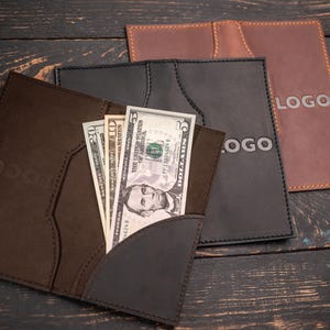 May include: Three leather wallets in brown, gray, and tan, with a dark wood background. The front wallet holds several US dollar bills. Each wallet has a stitched border and a logo.