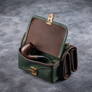 Puede incluir: Un estuche de cuero verde para pipa con interior marr&oacute;n y una pipa de madera en su interior. El estuche tiene forma rectangular con tapa abisagrada y cierre dorado. El estuche est&aacute; sobre una superficie gris.