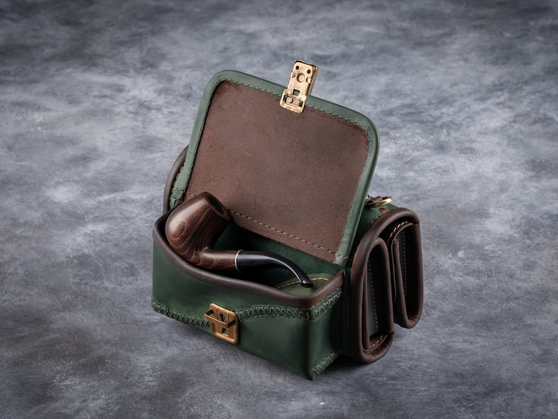 Puede incluir: Un estuche de cuero verde para pipa con interior marr&oacute;n y una pipa de madera en su interior. El estuche tiene forma rectangular con tapa abisagrada y cierre dorado. El estuche est&aacute; sobre una superficie gris.
