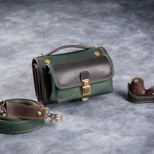 Puede incluir: Un bolso de cuero verde y marr&oacute;n con una correa para el hombro a juego y una pipa de madera. El bolso tiene un asa superior y una solapa frontal. La pipa descansa sobre un soporte de cuero. Los art&iacute;culos est&aacute;n sobre una superficie gris.