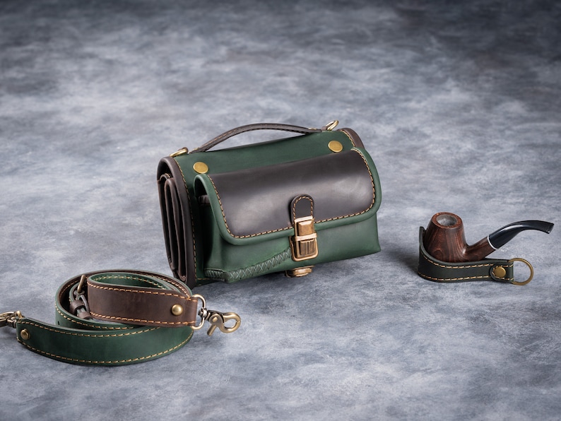 Puede incluir: Un bolso de cuero verde y marr&oacute;n con una correa para el hombro a juego y una pipa de madera. El bolso tiene un asa superior y una solapa frontal. La pipa descansa sobre un soporte de cuero. Los art&iacute;culos est&aacute;n sobre una superficie gris.