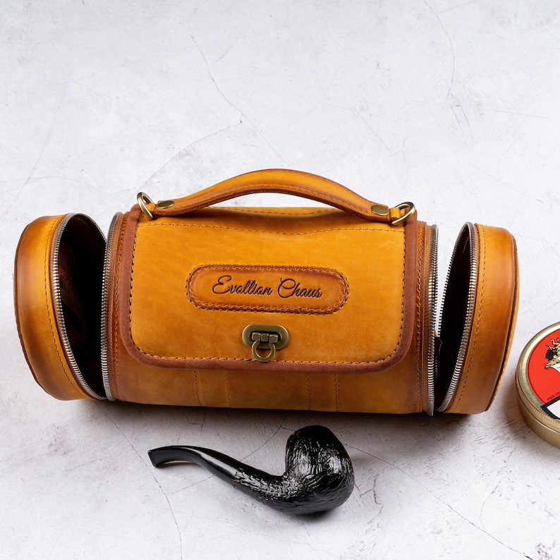 May include: A brown leather travel bag with a zippered opening and a brass clasp. The bag has the text "Evellion Chaus" embossed on the front. A black pipe is in the foreground.