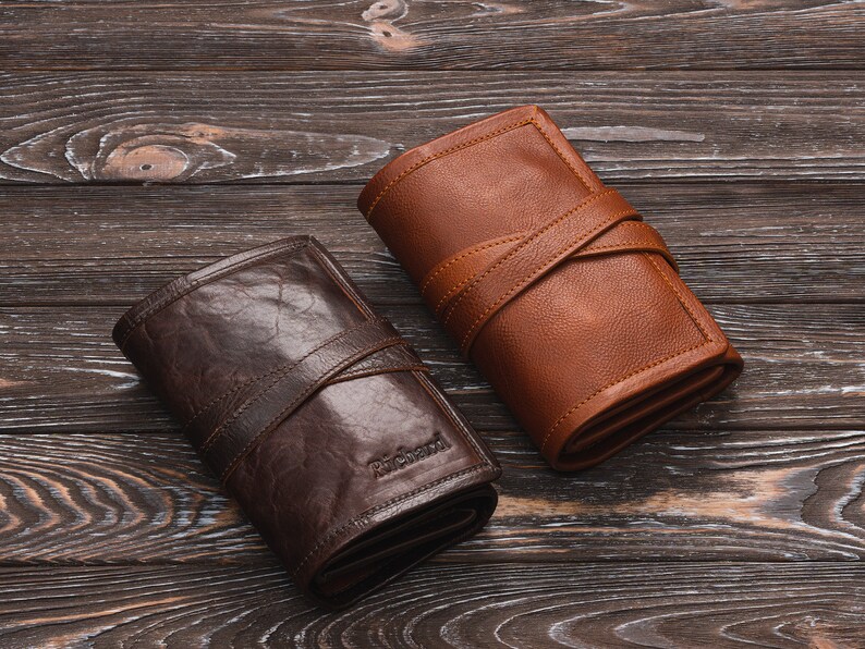 May include: Two leather wallets, one brown and one dark brown, with a strap closure. The wallets are on a wooden surface.