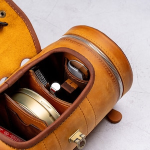 May include: A brown leather travel case with a zipper closure and multiple compartments. The case is open to reveal a small round container, a metal tool, and a brown leather pouch.