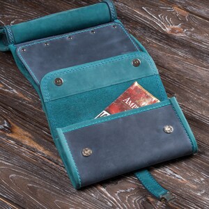 Puede incluir: Estuche enrollable de cuero artesanal en azul verdoso y azul marino. El estuche est&aacute; abierto, revelando bolsillos interiores y un libro parcialmente visible con la palabra "AMP" en la portada. El estuche se asegura con cierres de presi&oacute;n y una correa.