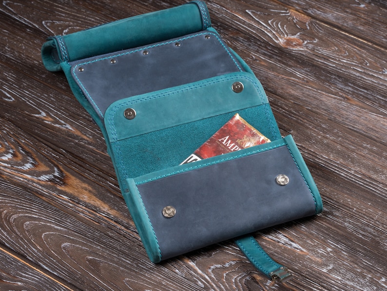 Puede incluir: Estuche enrollable de cuero artesanal en azul verdoso y azul marino. El estuche est&aacute; abierto, revelando bolsillos interiores y un libro parcialmente visible con la palabra "AMP" en la portada. El estuche se asegura con cierres de presi&oacute;n y una correa.