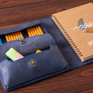 May include: A blue leather notebook with a zippered pocket for pencils and a separate pocket for a notepad. The notepad has a brown cover with a drawing of a fish and the words "Dream Big".