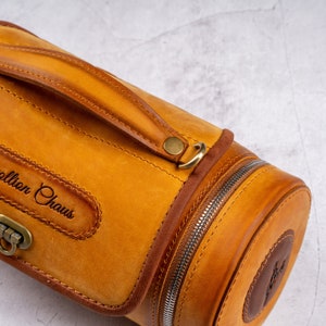 May include: A cylindrical leather travel bag with a brown leather handle and a zipper closure. The bag has a brass buckle and a leather patch with the text "Collion Chaus" embossed on it.
