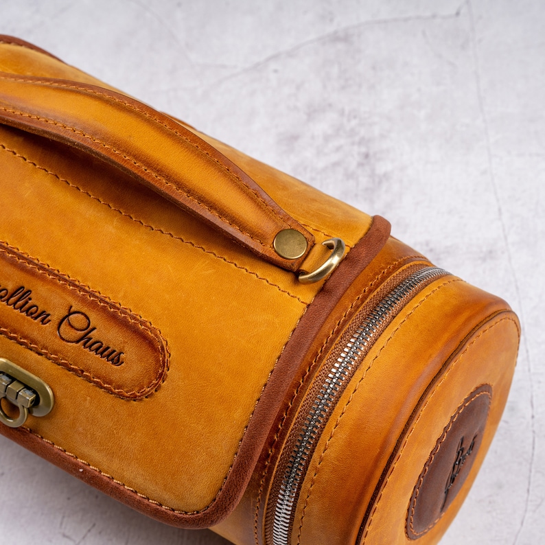 May include: A cylindrical leather travel bag with a brown leather handle and a zipper closure. The bag has a brass buckle and a leather patch with the text "Collion Chaus" embossed on it.