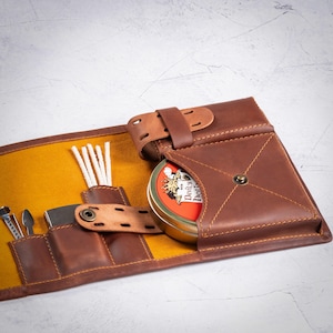 May include: A brown leather tobacco pouch with a yellow interior, holding a tin of tobacco, cleaning tools, and pipe cleaners. The pouch is secured with a leather strap and buckle, and features stitched detailing.