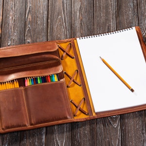 May include: A brown leather sketchbook with a built-in pencil holder. The sketchbook has a yellow leather flap with a criss-cross lacing detail. The sketchbook is open to reveal a blank white page and a yellow pencil.
