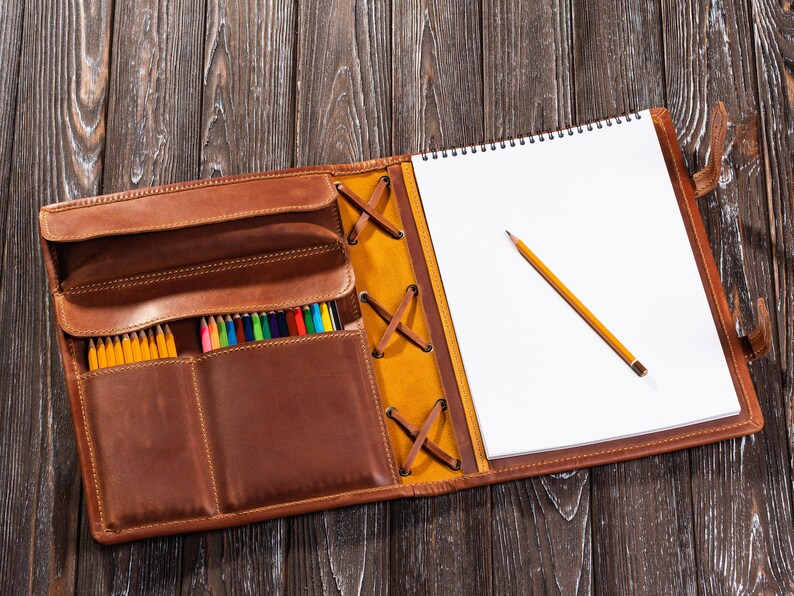 May include: A brown leather sketchbook with a built-in pencil holder. The sketchbook has a yellow leather flap with a criss-cross lacing detail. The sketchbook is open to reveal a blank white page and a yellow pencil.