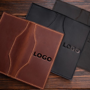 May include: Three leather items, including a brown wallet with the word "LOGO" embossed on the front. Two black wallets are also visible, each with the word "LOGO" embossed. All items are on a wooden surface.