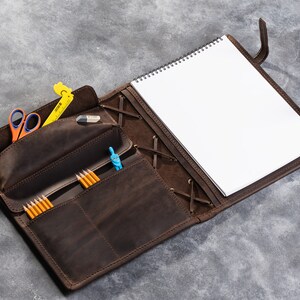 May include: A brown leather portfolio with a notebook, pencils, a ruler, a compass, and scissors. The portfolio has a flap that closes with a strap and buckle.