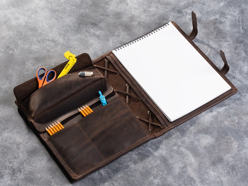 May include: A brown leather portfolio with a notebook, pencils, a ruler, a compass, and scissors. The portfolio has a flap that closes with a strap and buckle.