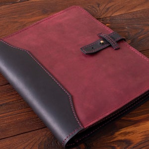 May include: A burgundy leather folder with a black leather flap and a strap closure. The folder has a stitched border and is lying on a wooden surface.