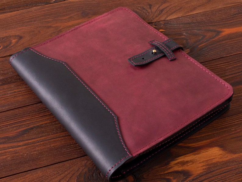 May include: A burgundy leather folder with a black leather flap and a strap closure. The folder has a stitched border and is lying on a wooden surface.