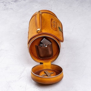 May include: A tan leather cylindrical travel case with a zipper closure and a small pocket inside. The case is open and the inside is visible, revealing a brown leather pocket and a silver lighter.