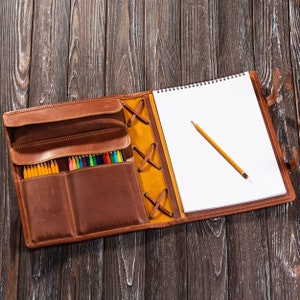 May include: A brown leather portfolio with a notepad and a set of colored pencils. The portfolio has a flap with a yellow and brown strap and a zipper closure. The notepad has a spiral binding and is open to a blank page. A yellow pencil is lying on the page.