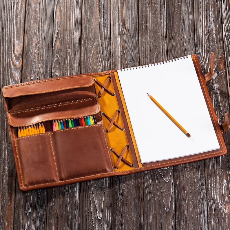 May include: A brown leather portfolio with a notepad and a set of colored pencils. The portfolio has a flap with a yellow and brown strap and a zipper closure. The notepad has a spiral binding and is open to a blank page. A yellow pencil is lying on the page.
