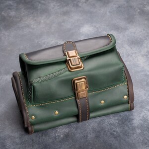 Puede incluir: Bolso de cuero artesanal en tonos verdes y marrones, con cierre de solapa y hebilla de lat&oacute;n. El bolso tiene costuras decorativas y remaches, con un dise&ntilde;o estructurado. El bolso est&aacute; dise&ntilde;ado para ser llevado.