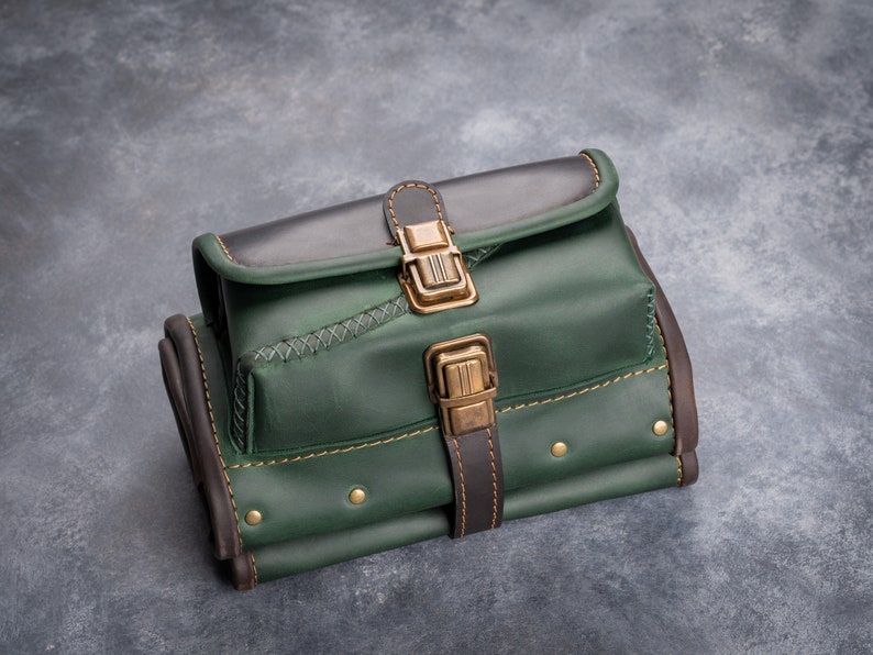 Puede incluir: Bolso de cuero artesanal en tonos verdes y marrones, con cierre de solapa y hebilla de lat&oacute;n. El bolso tiene costuras decorativas y remaches, con un dise&ntilde;o estructurado. El bolso est&aacute; dise&ntilde;ado para ser llevado.