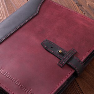 May include: A burgundy leather journal with a black leather flap and a black leather strap with a brass buckle. The journal has a red stitching detail and the text "E. Bondarenko" embossed on the front cover.