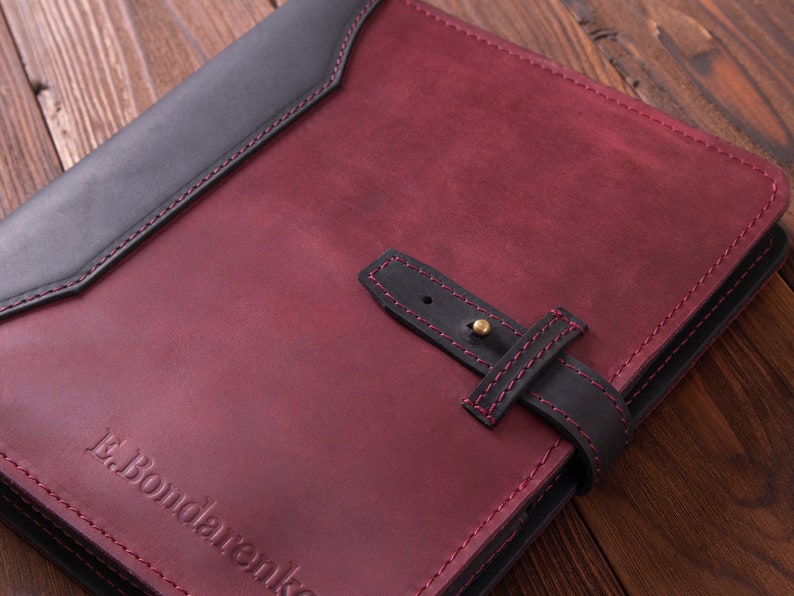 May include: A burgundy leather journal with a black leather flap and a black leather strap with a brass buckle. The journal has a red stitching detail and the text "E. Bondarenko" embossed on the front cover.