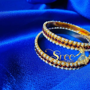 May include: Two gold bangles with red and white beads. The bangles are stacked on top of each other and are on a blue satin background.