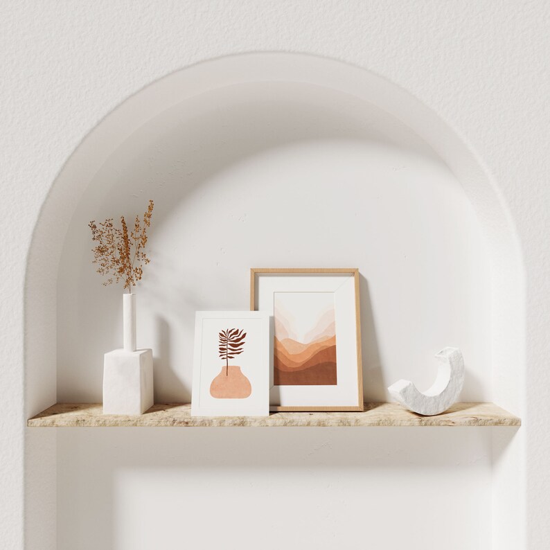 May include: Two framed prints with abstract designs in shades of brown and pink, a white vase with dried flowers, and a white decorative object on a wooden shelf in a white arched alcove.