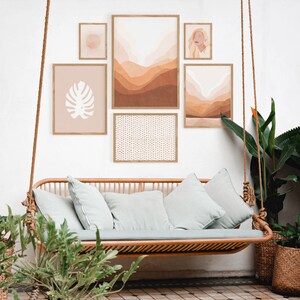 May include: A light brown wicker swing sofa with light blue cushions, hanging from a white wall with a collection of framed artwork. The artwork includes a brown and white abstract landscape, a white silhouette of a monstera leaf, a white and brown geometric pattern, and a portrait of a woman with long blonde hair.