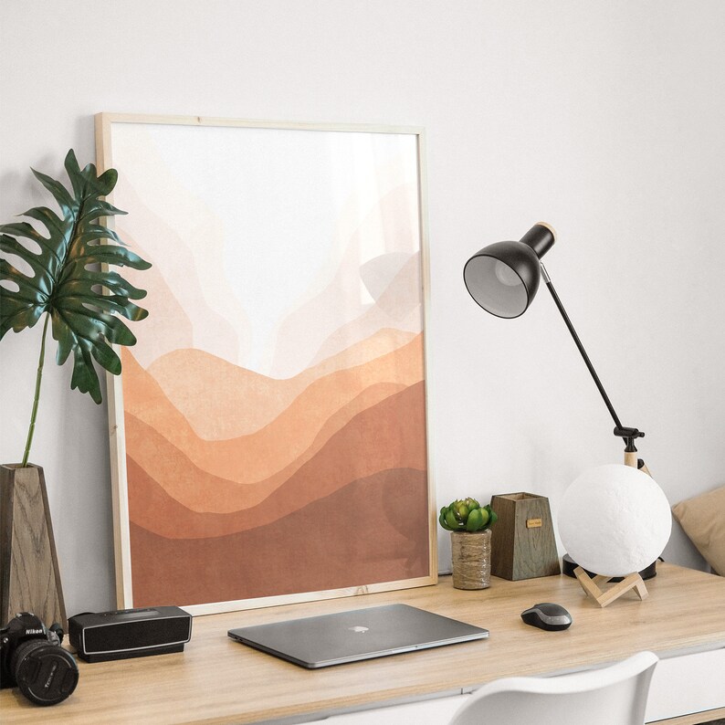 May include: A framed abstract print with a watercolor-like design of brown, orange, and beige hills. The print is hanging on a white wall above a desk with a laptop, a lamp, and a small potted plant.