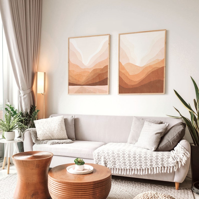 May include: A light beige sofa with two throw pillows and a throw blanket in a living room setting. Two framed abstract landscape prints with brown and beige tones hang on the wall above the sofa. A wooden coffee table sits in front of the sofa.