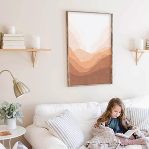 May include: A framed abstract print with a landscape of brown, orange, and pink watercolor hills. The print is hanging on a white wall above a white couch with a girl sitting on it reading a book.