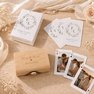 May include: A wooden box engraved with the names Mia & Noah and the date 06.14.2026, alongside a deck of playing cards in a white box and loose cards featuring a floral wreath design with the names Ethan & Lucy and the date 09.21.2026.