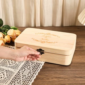 Peut inclure: Une boîte en bois de couleur claire avec un couvercle à charnière et un fermoir en métal foncé. Le couvercle est gravé des noms "Lelilia & Andre" et de motifs floraux. La boîte est rectangulaire et semble être en pin.