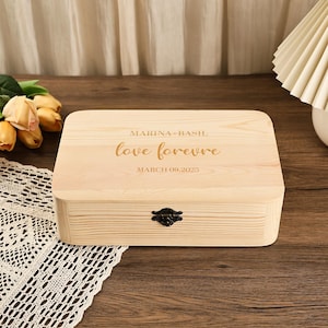 May include: A light-coloured wooden box with a hinged lid and a decorative metal clasp. The lid is engraved with the words "MARINA + BASIL", "love forever", and "MARCH 09,2025". The box is rectangular and appears to be made of pine.