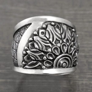 May include: A silver ring with a wide band featuring an intricate, detailed mandala design.
