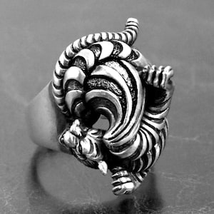 May include: A silver-colored ring featuring a detailed, three-dimensional tiger design. The tiger is depicted in a dynamic pose, with its body curved and its head and tail raised. The ring has a wide band and a textured surface, showcasing the tiger's stripes and features.