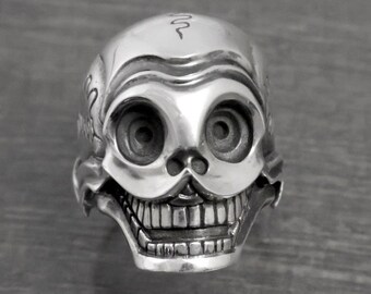 May include: A silver skull ring featuring a wide, toothy grin and large, round eyes. The skull is intricately designed with detailed lines and a small snake symbol on the forehead.
