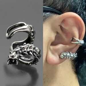 May include: Silver ear cuff in the shape of a dragon. The cuff is worn on the upper part of the ear and has a detailed design.