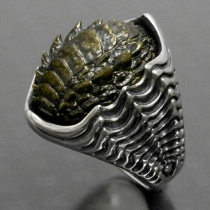 May include: A silver ring with a detailed, textured design. The ring features a bronze-colored, crustacean-like creature on top. The band has a ribbed pattern. The ring is set against a dark background.