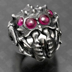 May include: A silver spider ring with three large, bright pink gemstones for eyes.