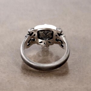 K&ouml;nnte beinhalten: Ein silberner Ring mit einem verzierten Design. Der Ring hat einen rechteckigen, wei&szlig;en Stein, der in einer detaillierten Filigranfassung gefasst ist. Das Band ist glatt und abgerundet, mit einer leicht oxidierten Oberfl&auml;che, und der Ring wird aus einem Winkel betrachtet.