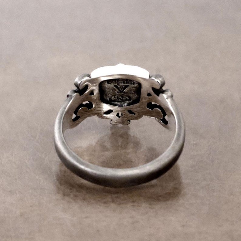 K&ouml;nnte beinhalten: Ein silberner Ring mit einem verzierten Design. Der Ring hat einen rechteckigen, wei&szlig;en Stein, der in einer detaillierten Filigranfassung gefasst ist. Das Band ist glatt und abgerundet, mit einer leicht oxidierten Oberfl&auml;che, und der Ring wird aus einem Winkel betrachtet.