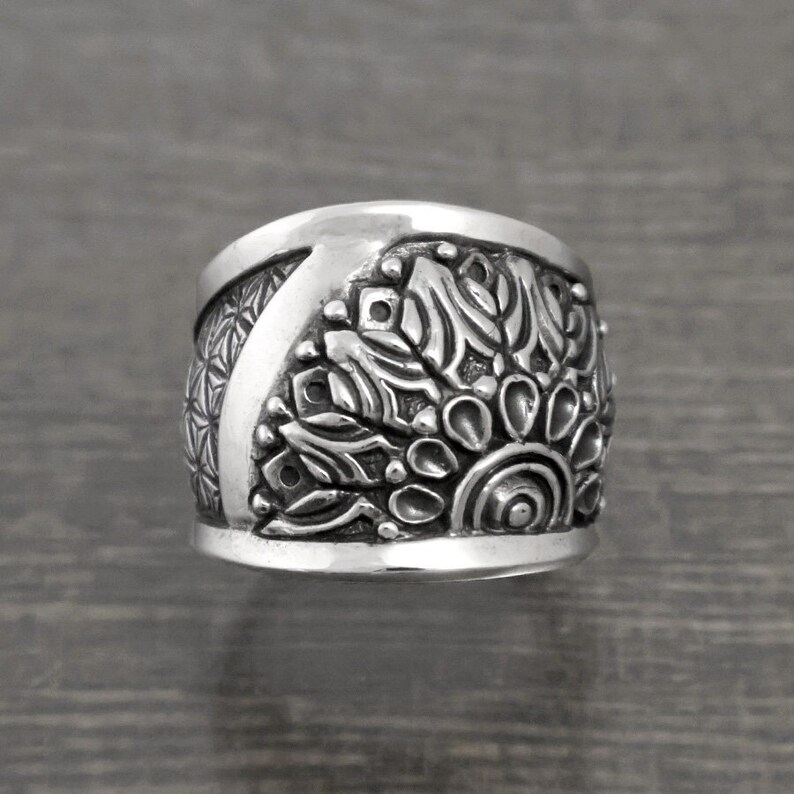 May include: A silver ring with a wide band featuring an intricate, detailed mandala design.